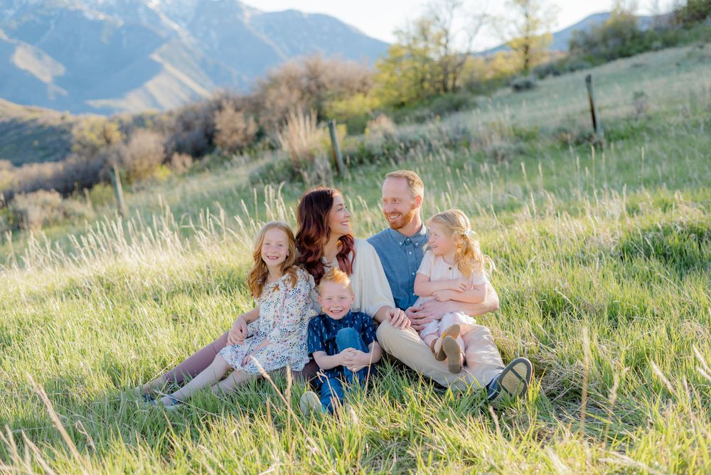 Family of five sitting together in a sunlit green meadow with rolling mountains in the background.