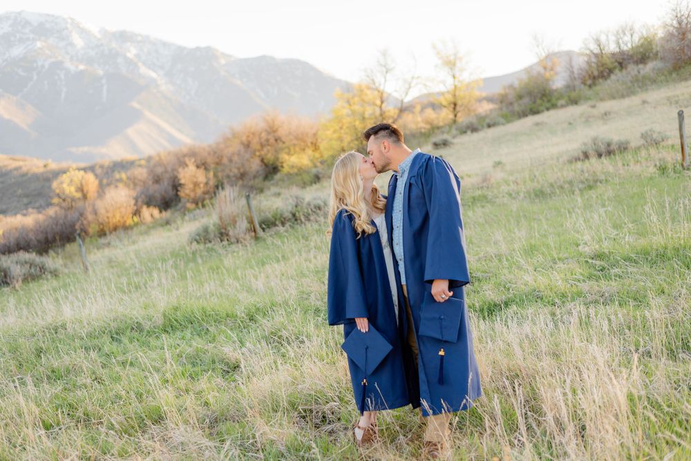 Couple sharing a kiss in graduation gowns on a green hillside with autumn trees and mountains in background.