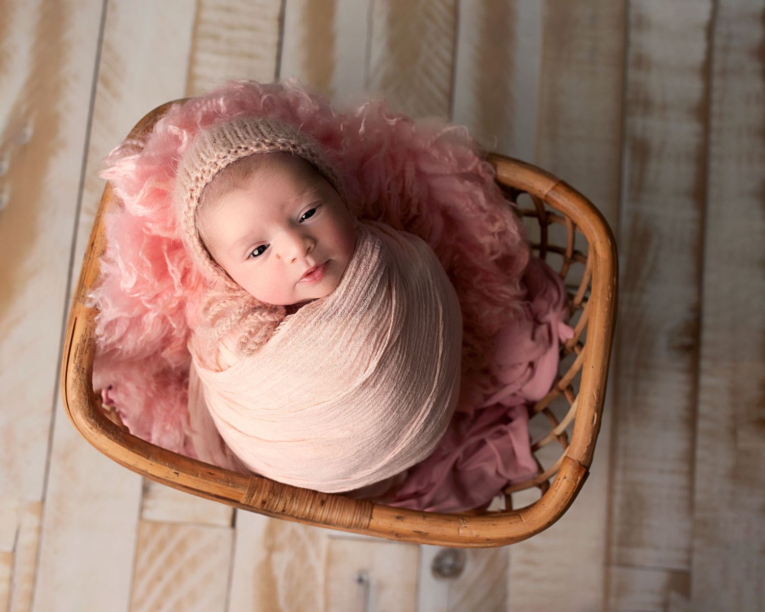Sleeping newborn baby wrapped in pink swaddle with knit bonnet nestled in a wicker basket.