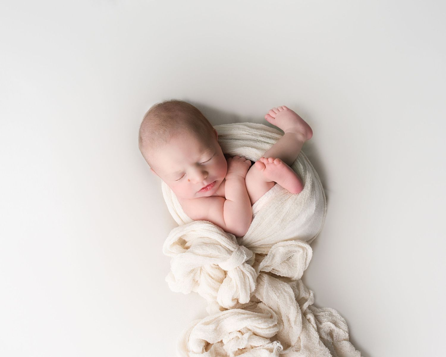 Peaceful newborn baby swaddled in cream white wrap sleeping on a soft white background.