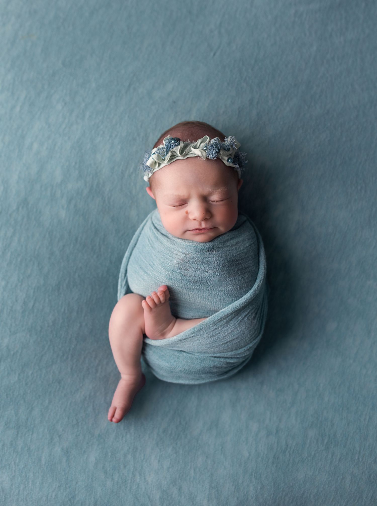 Newborn baby girl swaddled in teal wrap wearing floral headband, sleeping peacefully on teal backdrop.