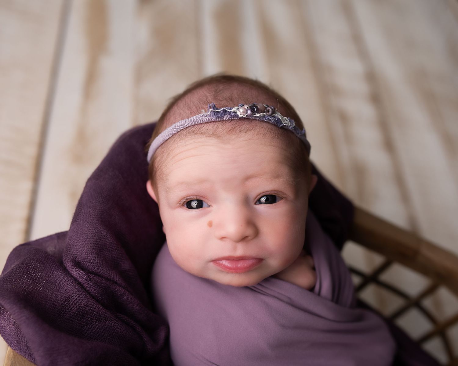 Alert newborn baby wrapped in purple swaddle wearing a silver headband in a wicker basket.