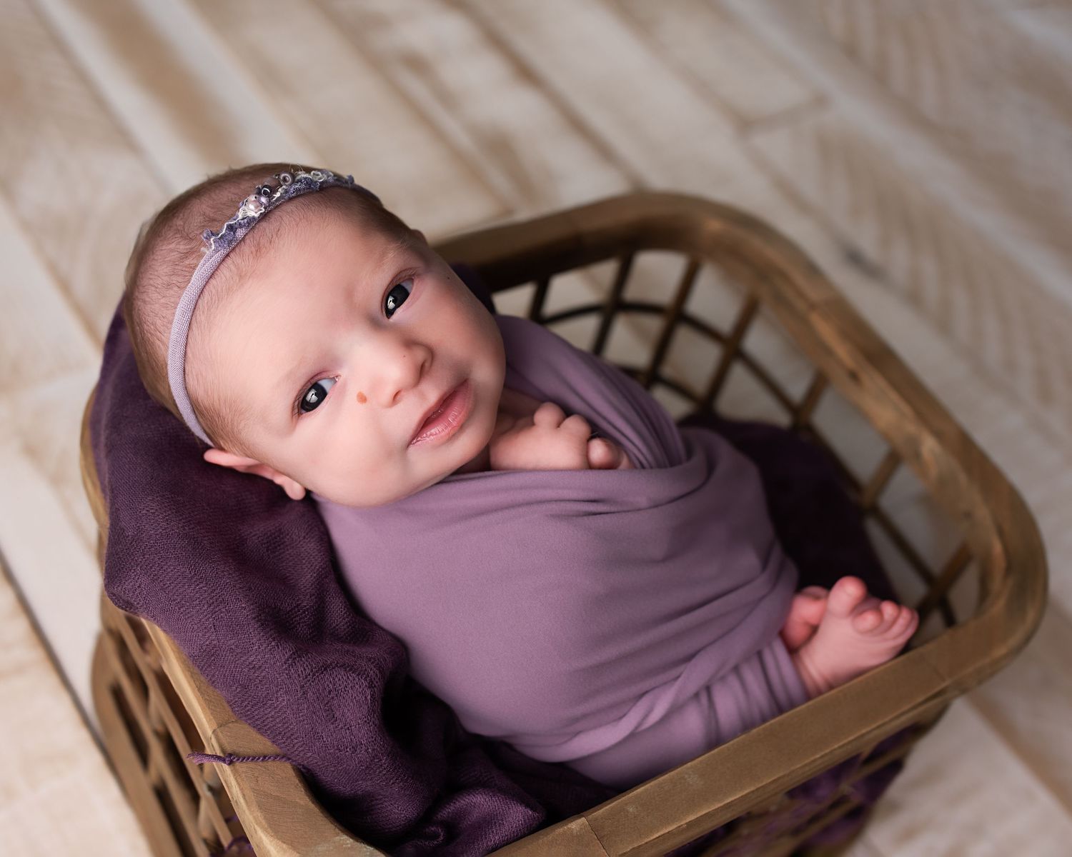 Wide-eyed newborn baby girl in purple wrap and silver headband posed in a wicker basket.