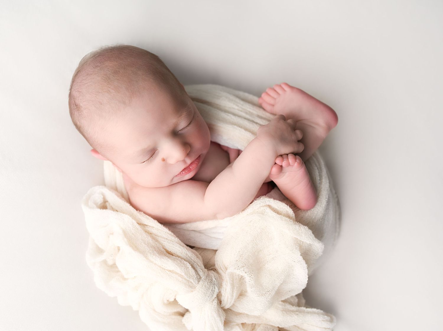 Sleeping newborn baby wrapped in cream fabric, curled peacefully in bowl on white background.