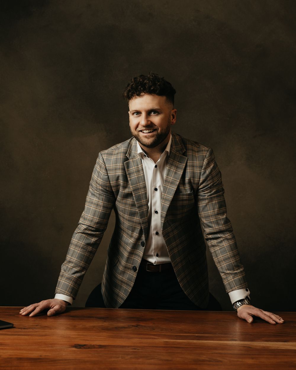 Business professional in plaid suit jacket leaning over wooden table in studio setting.