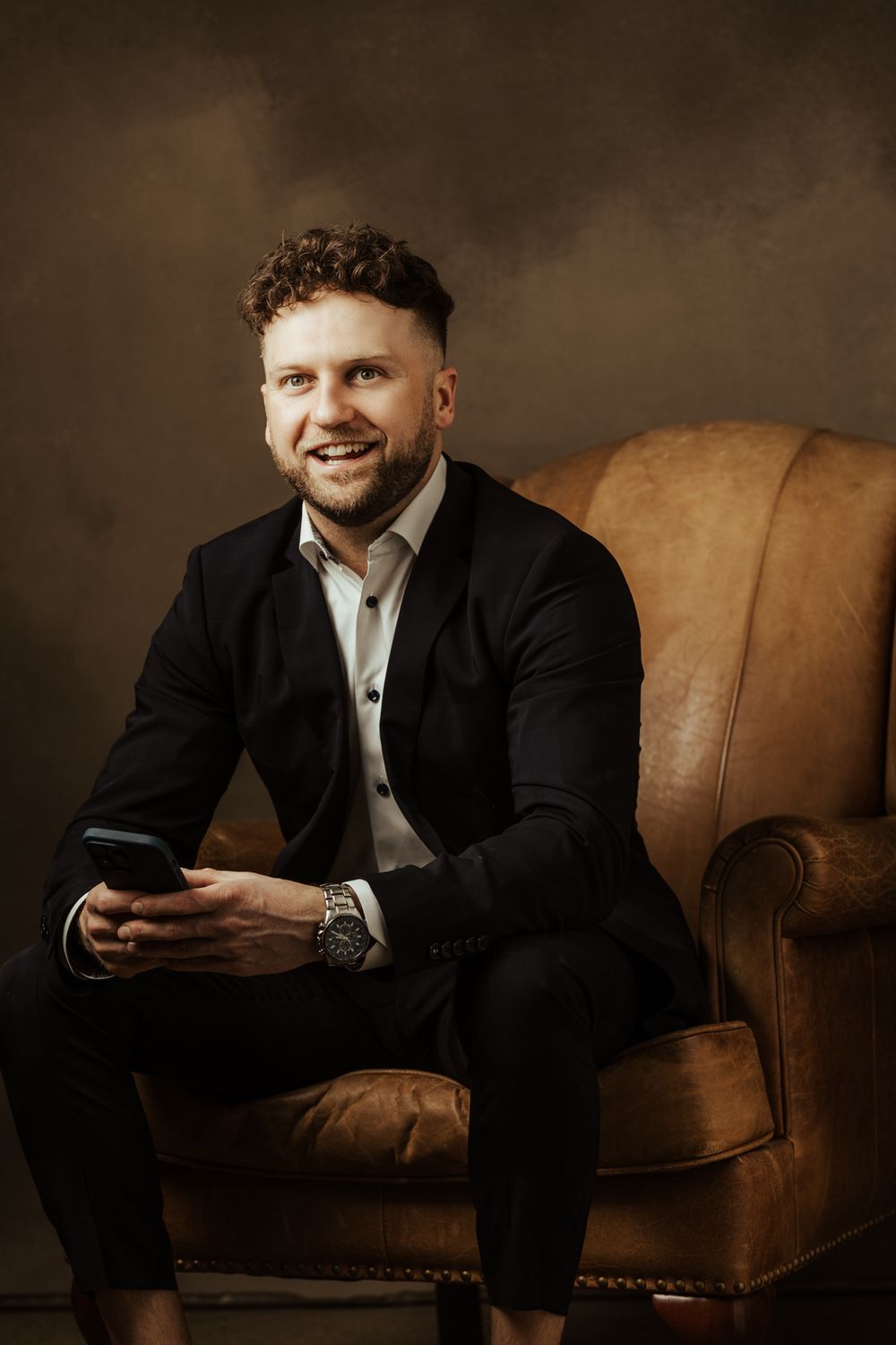 Professional portrait series of someone in a suit sitting on a brown leather couch against dark backdrop.