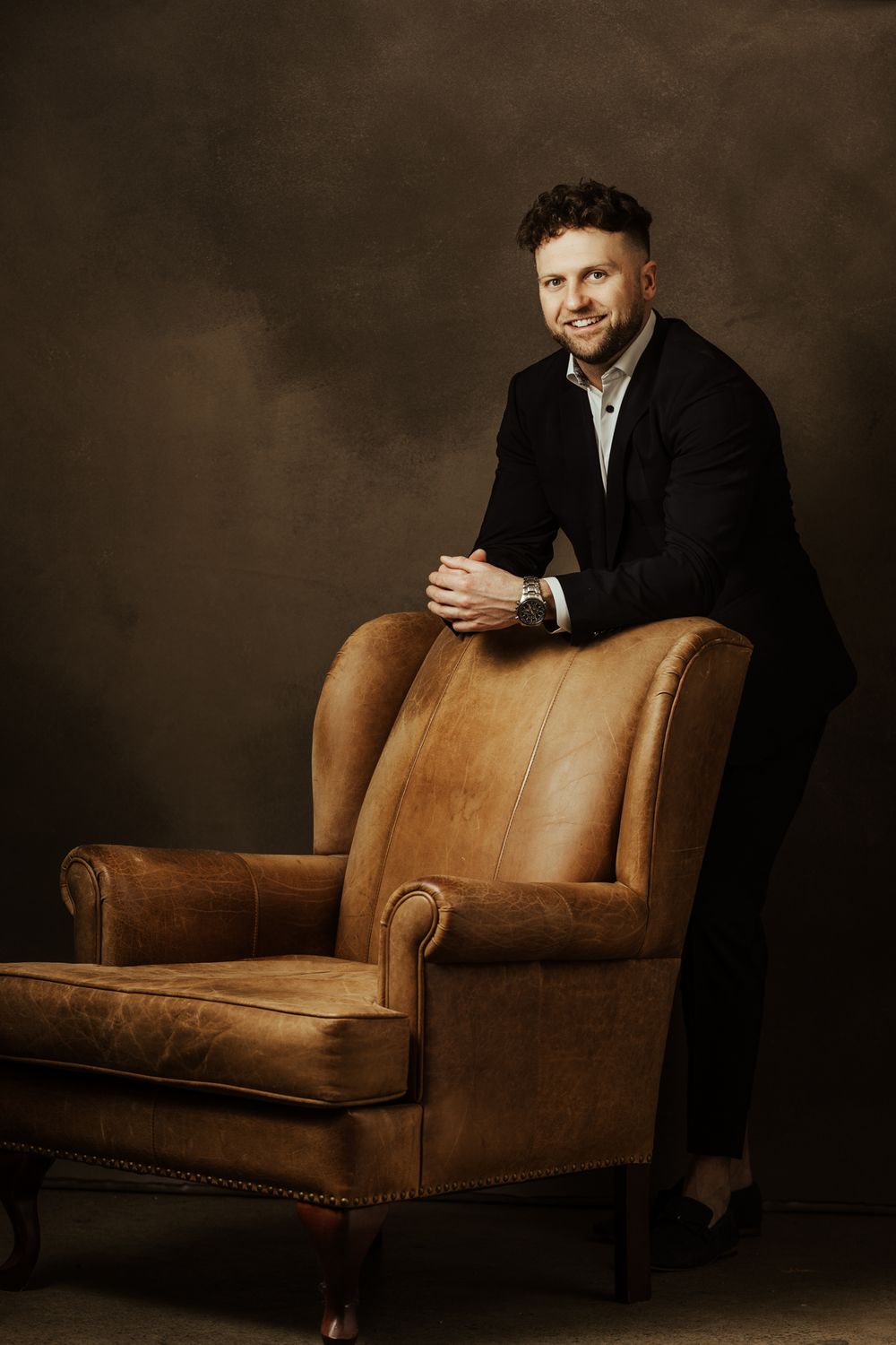 Moody portrait series showing someone in formal attire lounging on brown leather seating.