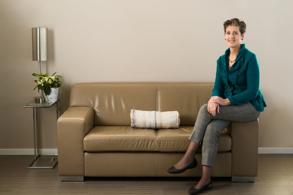 Person in teal blazer sits on beige couch in minimalist room with white walls and decorative plant.