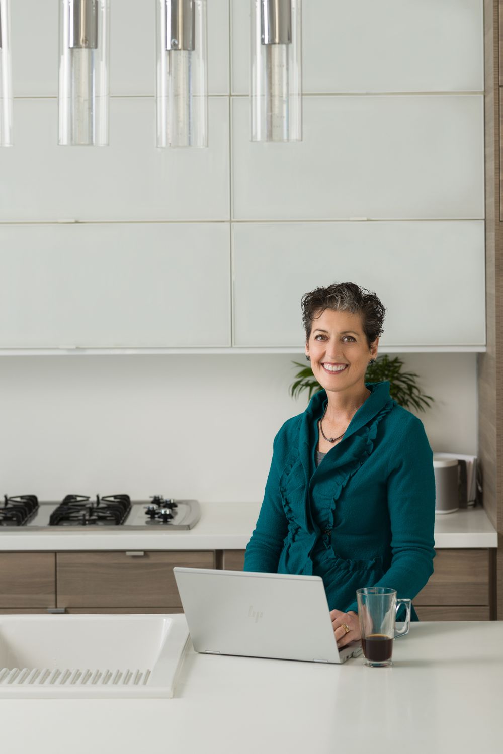 Person in teal jacket works at modern white kitchen counter with laptop.