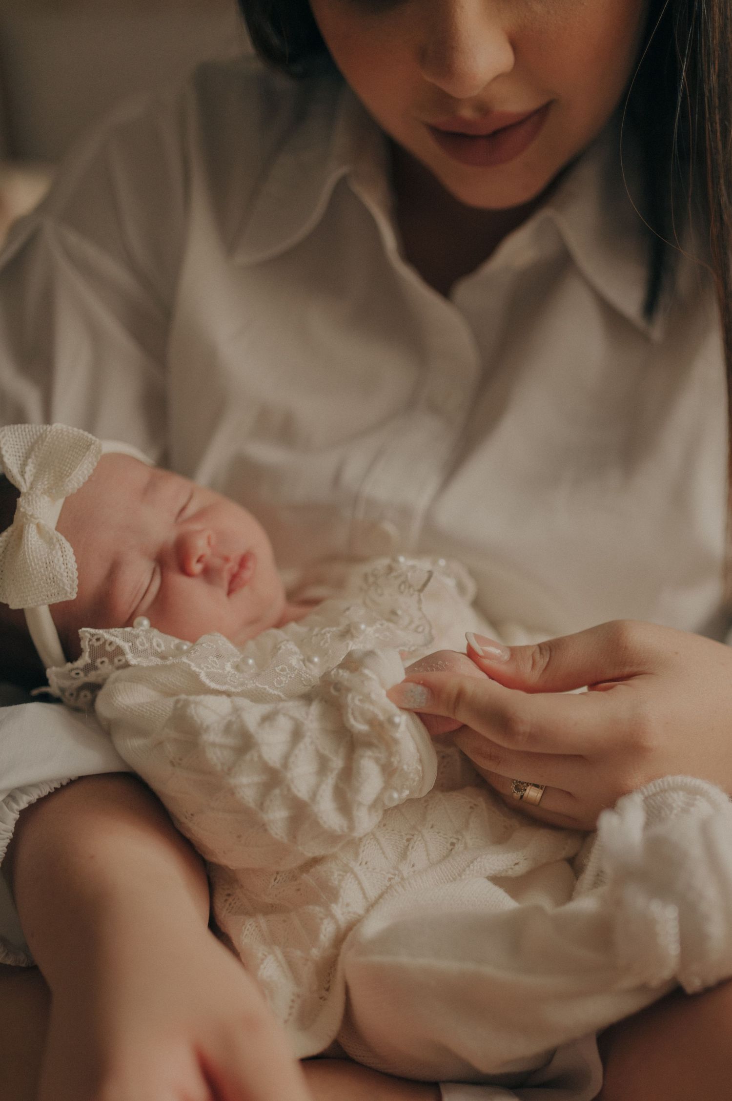 A sleeping newborn baby wearing a white lace dress and bow headband being gently held.