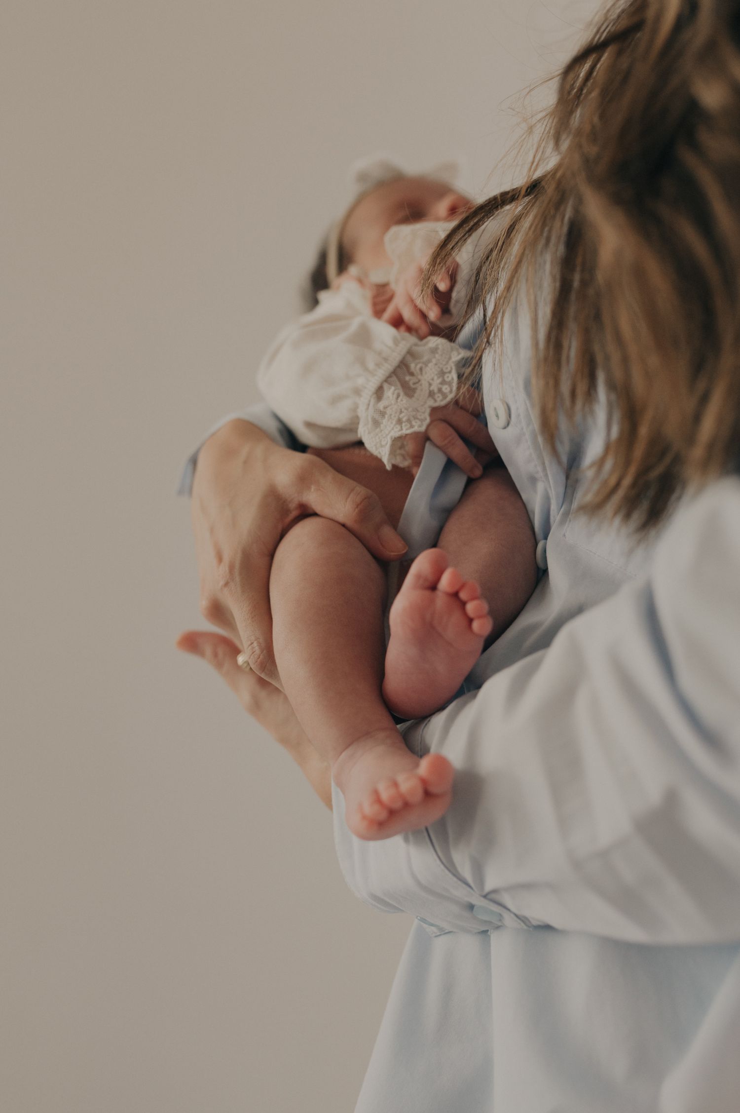 Tender portrait of parent holding baby in white lace outfit against neutral background.