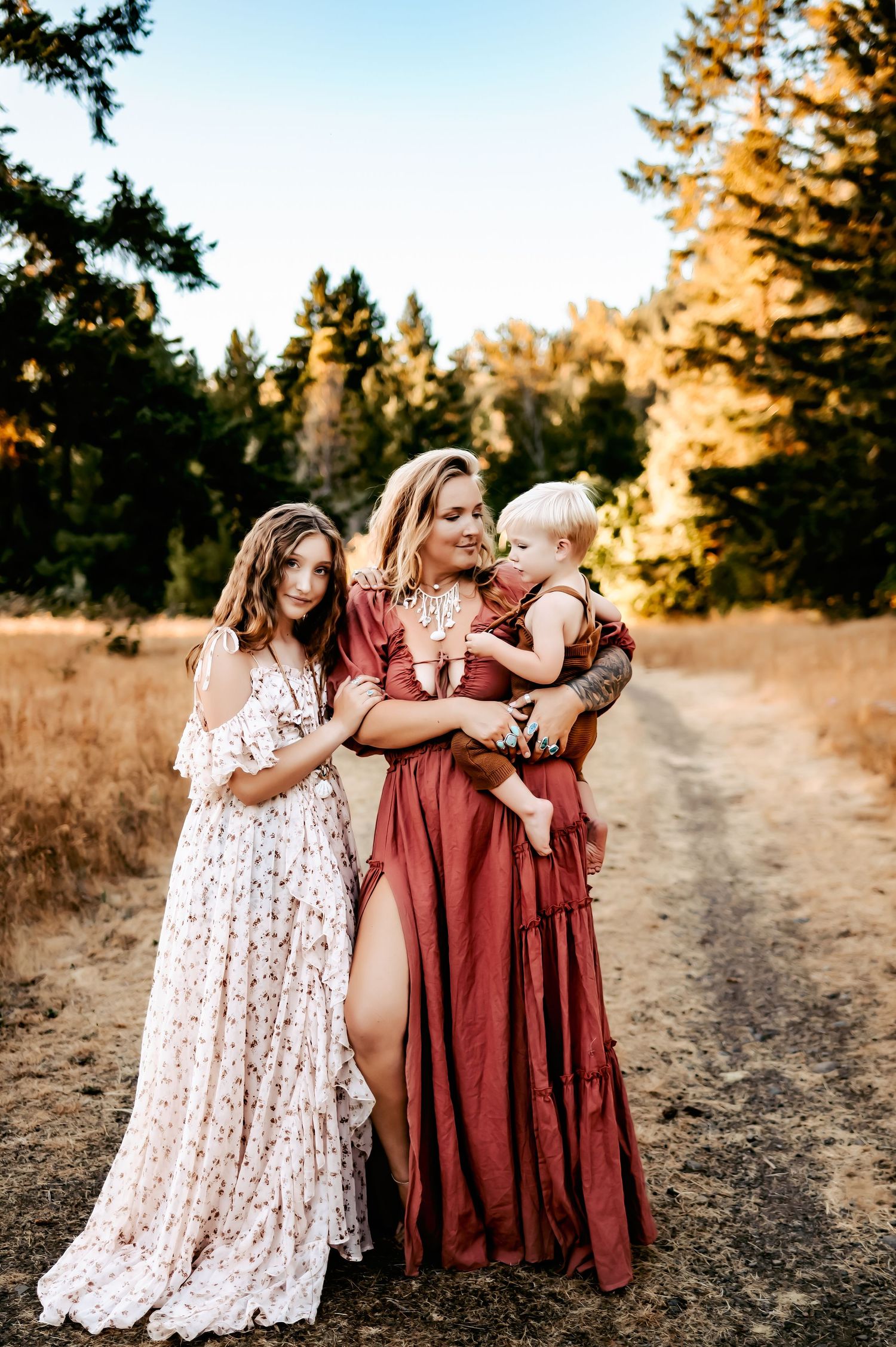 Three women in floral and burgundy dresses share an intimate moment on outdoor path.