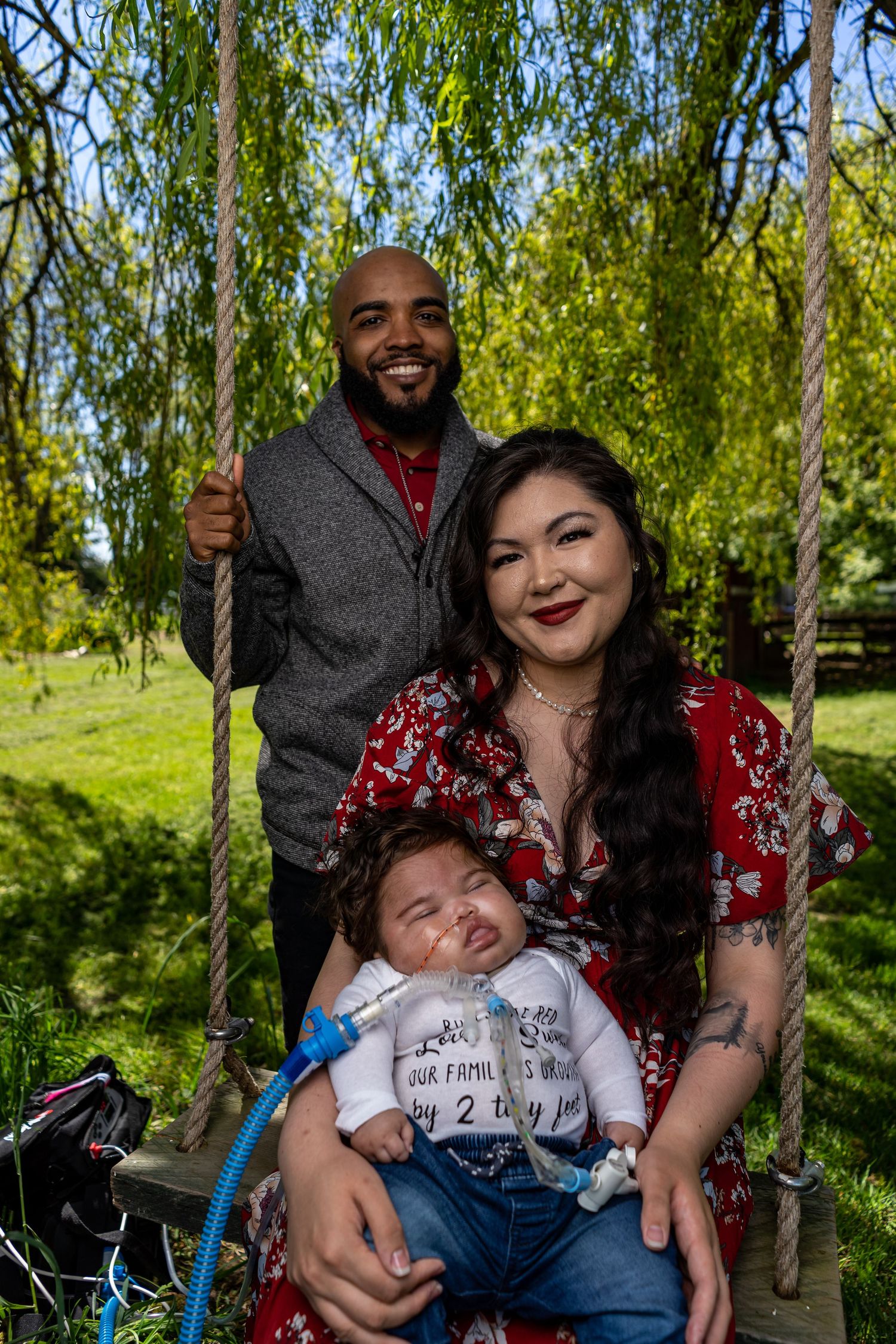 A family poses together on a swing under a weeping willow tree on a sunny day.