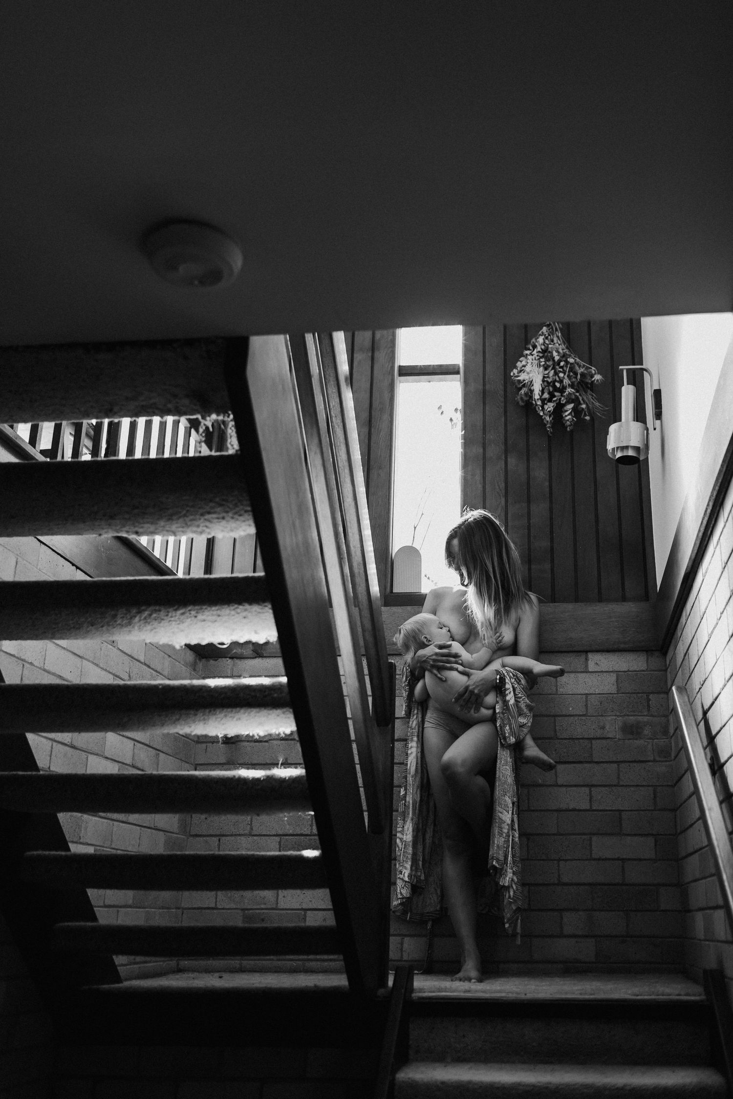Mother nursing newborn baby on staircase in moody black and white lifestyle photo.