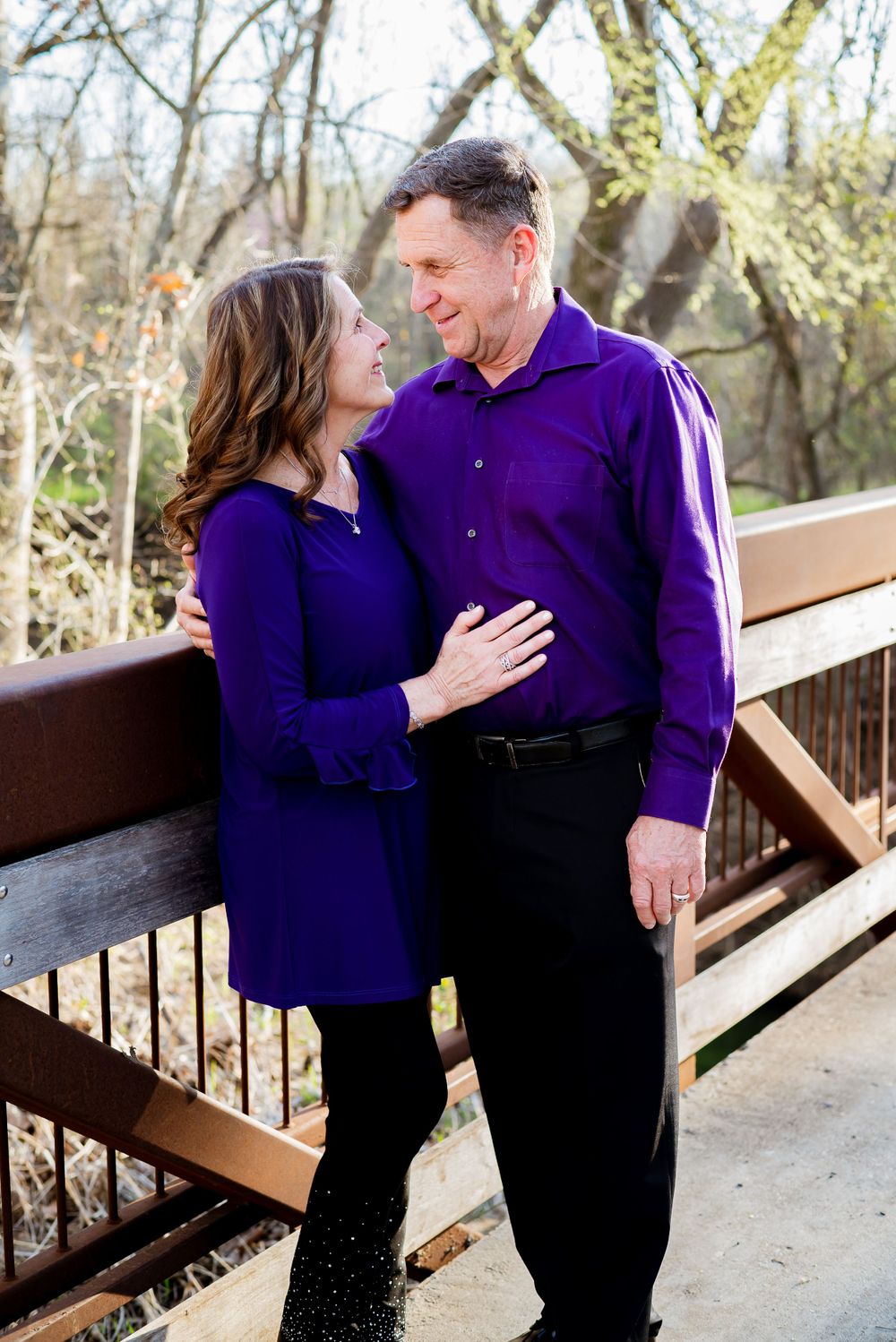 A couple wearing matching purple outfits share a tender moment on a wooden bridge.