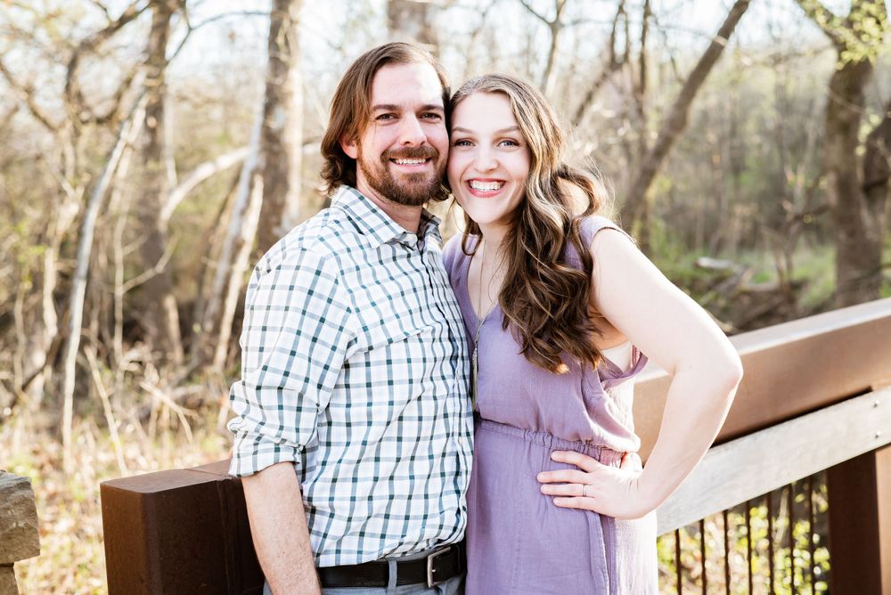 A couple poses together outdoors in a wooded area wearing coordinated casual outfits.