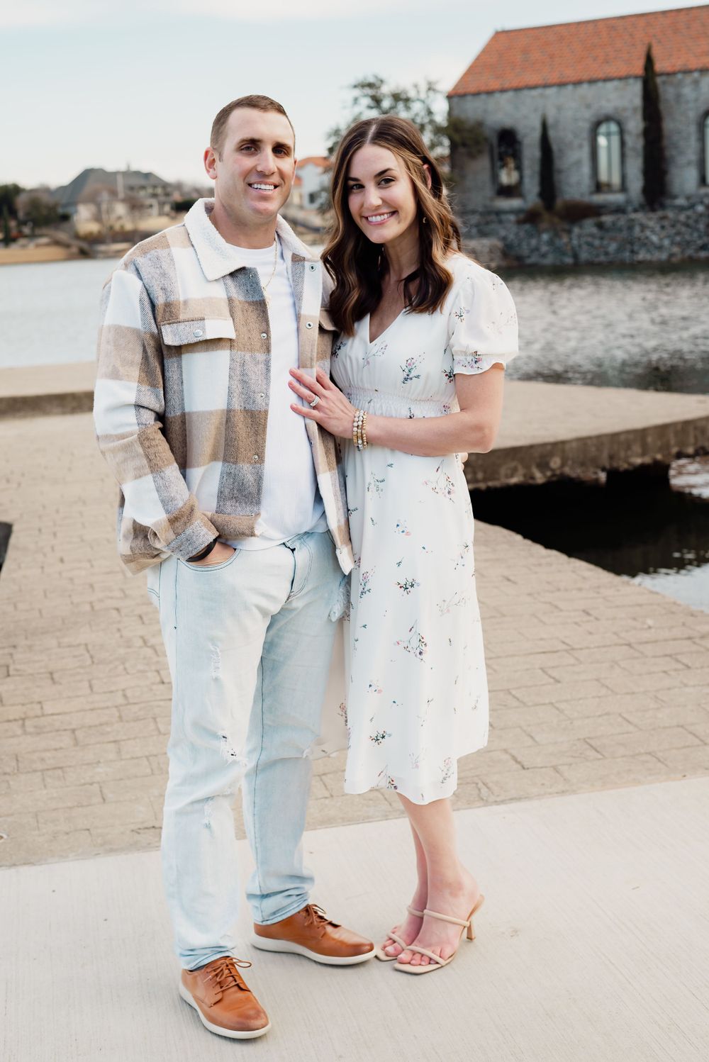 A couple poses together on a waterfront path with a historic building in the background.