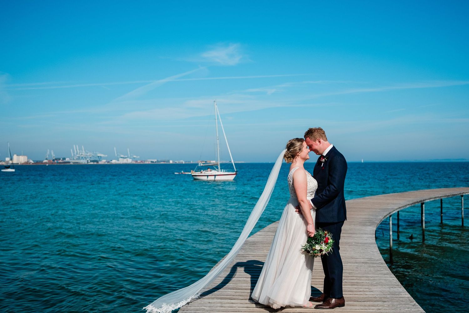 Couple sharing romantic moment on oceanfront pier with sailboat and blue waters in background.