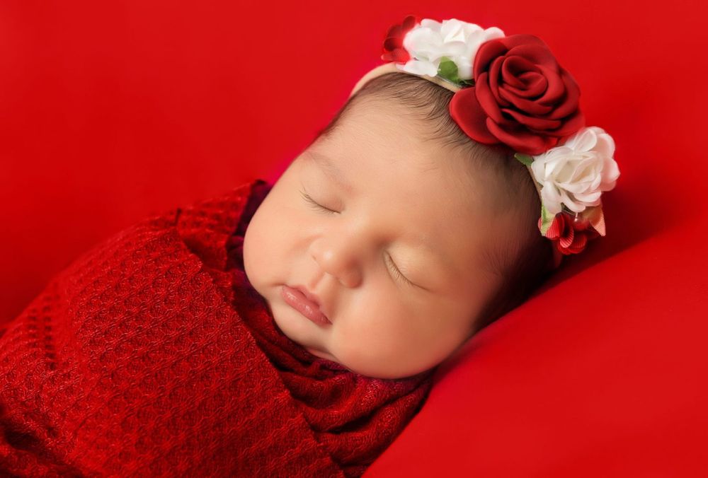 Sleeping newborn in red wrap with floral headband against bright red background.