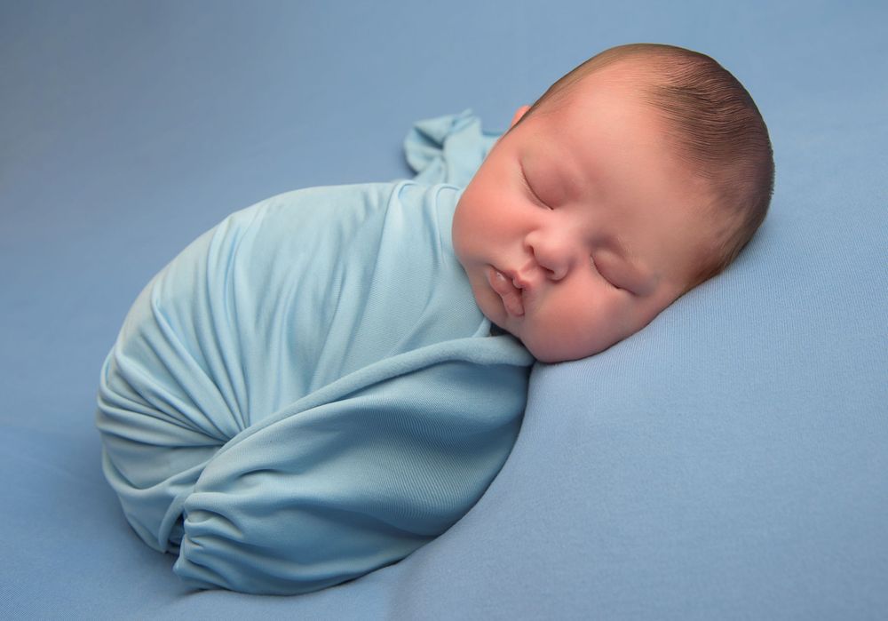 Peaceful sleeping newborn swaddled in light blue wrap against blue background.