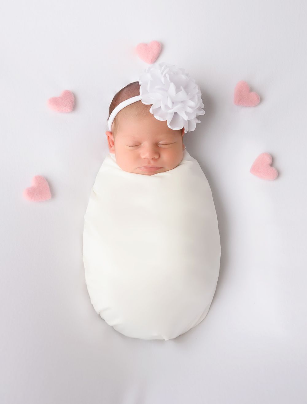 Swaddled newborn with white headband surrounded by pink hearts.