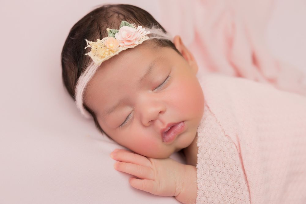 Sleeping newborn wearing lace headband and pink wrap on soft pink background.