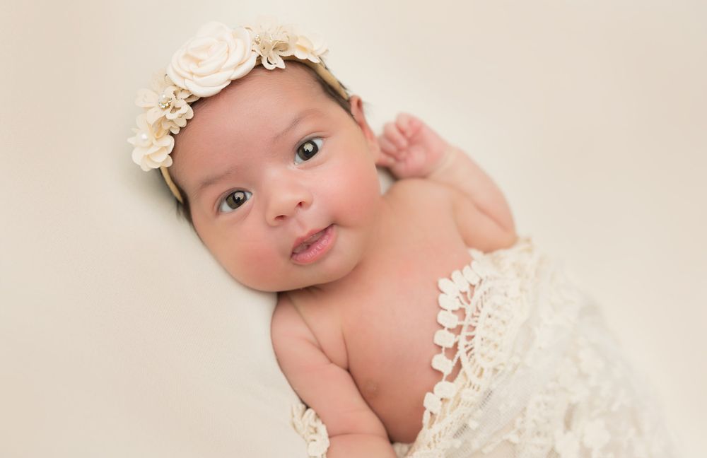 Alert newborn in delicate lace wrap and floral crown on pink background.