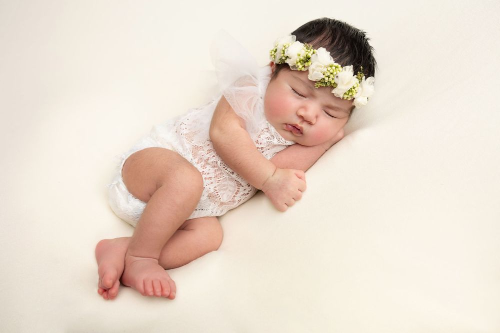 Sleeping baby wearing white lace romper and floral crown on cream background.