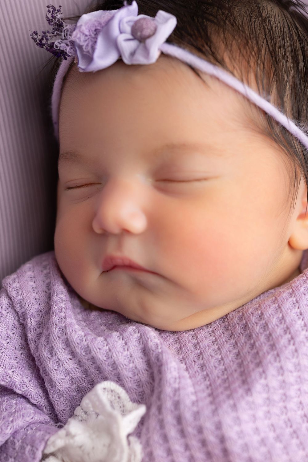 Sleeping baby wearing purple knit sweater and matching headband with floral detail.