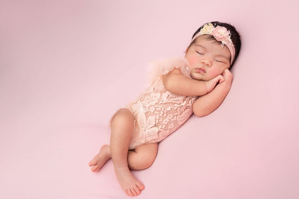 Sleeping newborn baby wearing pink lace romper on pink background during professional photo session.