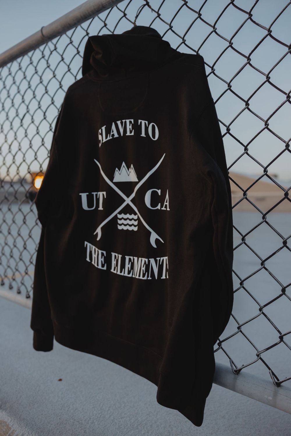Black hoodie with 'Slave to the Element' crossed arrows design displayed on chain link fence.
