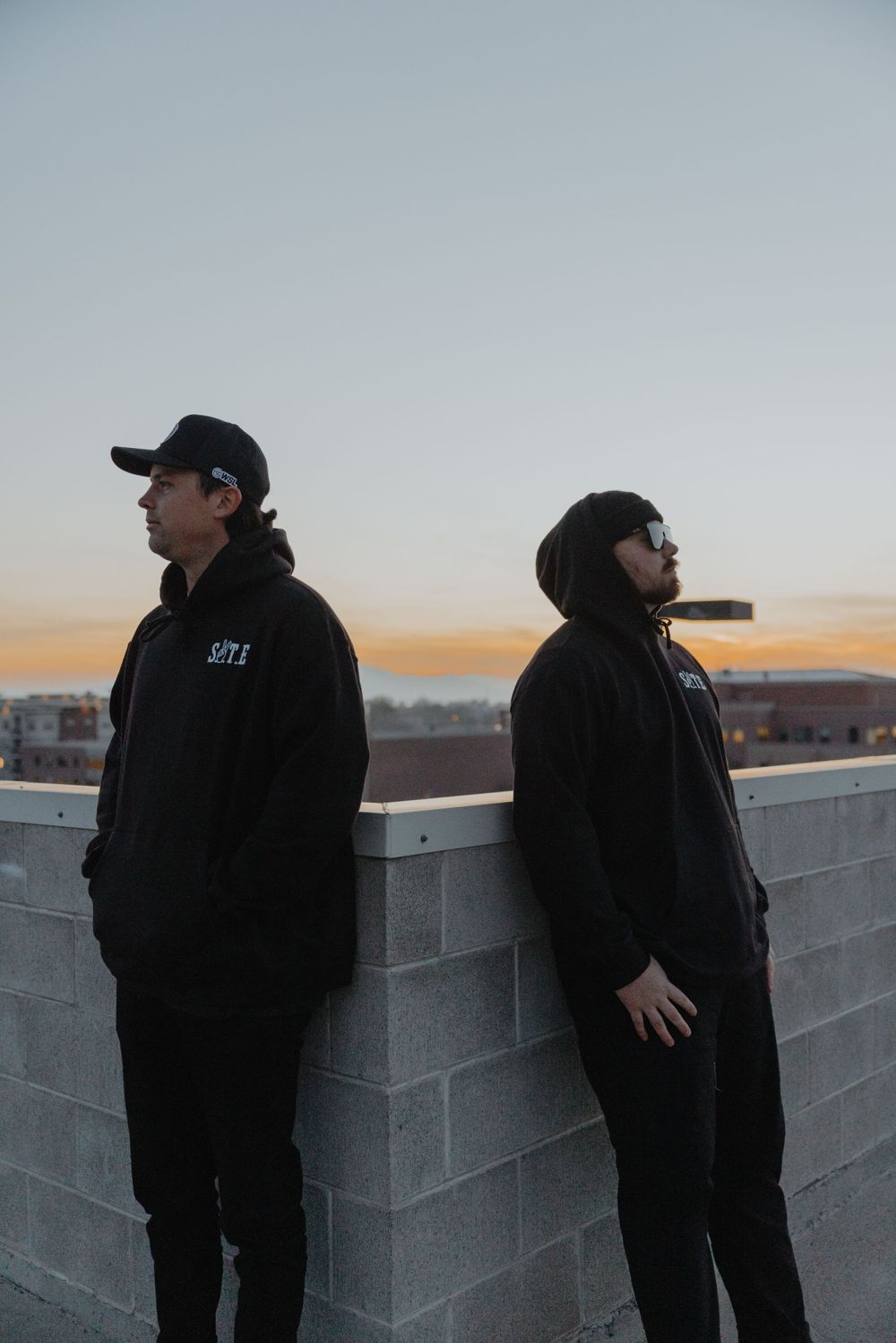 Two people in dark clothing standing against wall during sunset.