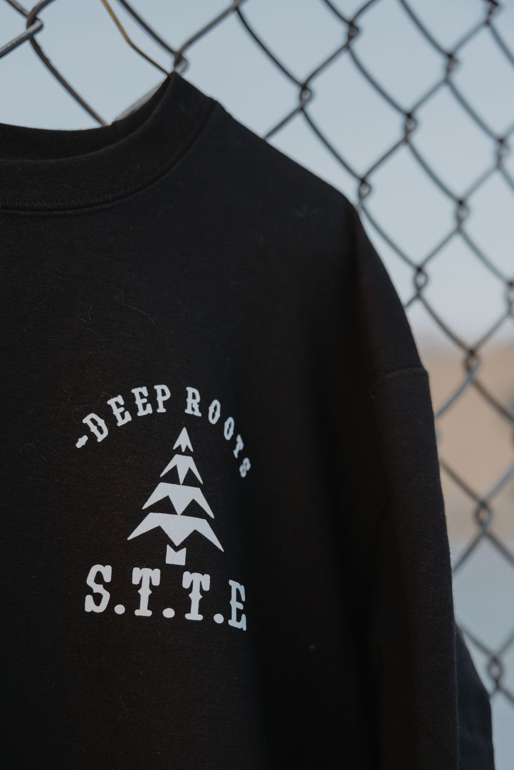 Black sweatshirt with Deep Roots STTE logo and mountain design hanging against chain link fence.