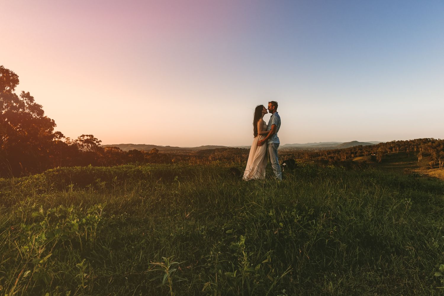 Romantic silhouette of couple standing in field during sunset with rolling hills in background.