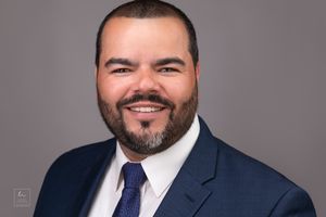 Professional headshot of person in navy suit and tie against grey background.
