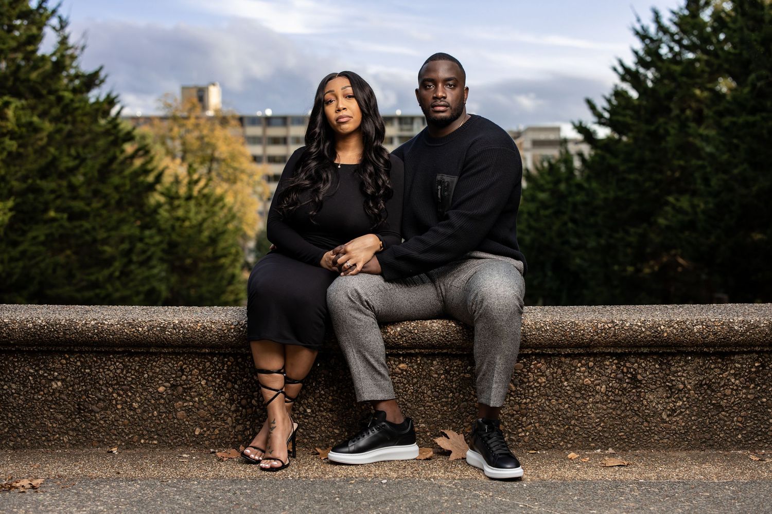 Couple in matching black outfits sitting together on stone wall with urban backdrop.