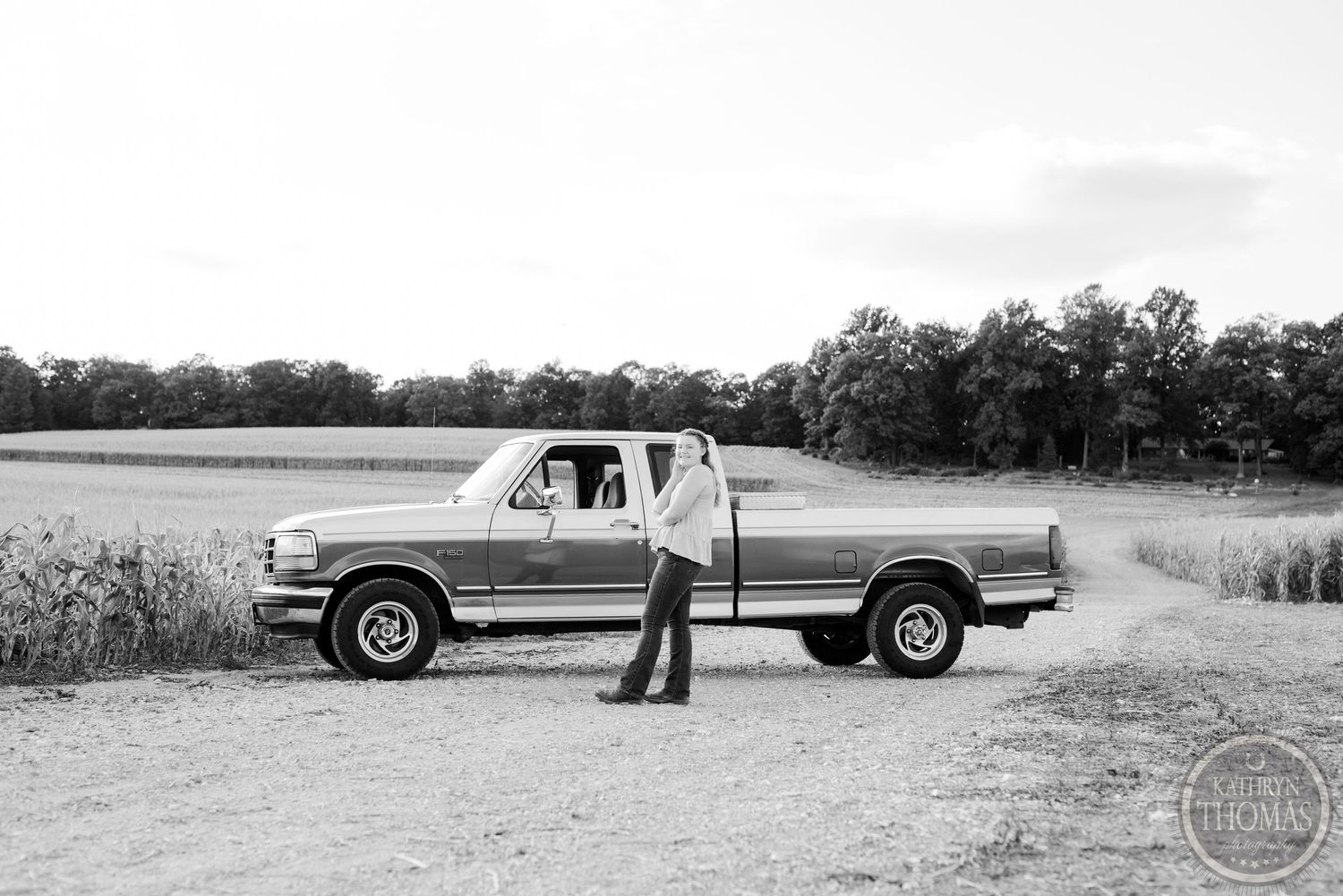 Black and white photo of a vintage pickup truck parked on a dirt road with fields in the background.
