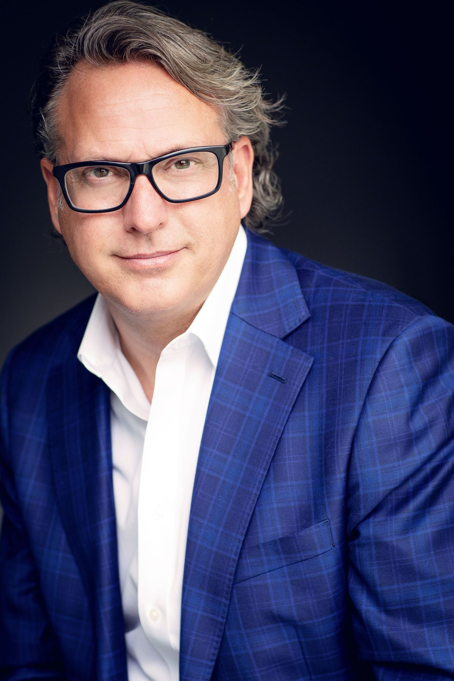 Executive portrait of person in blue checked blazer and glasses against dark backdrop.