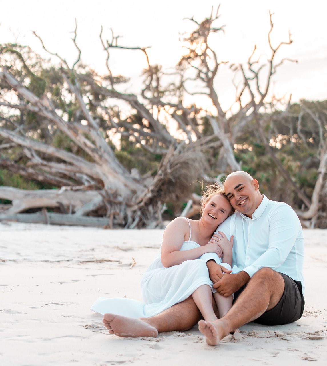 Couple relaxing on beach with driftwood and trees in background during golden hour.