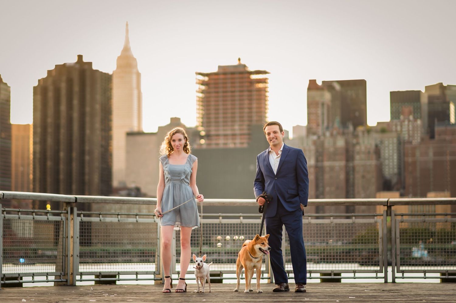 Couple with dog poses on waterfront walkway with Manhattan skyline in background.