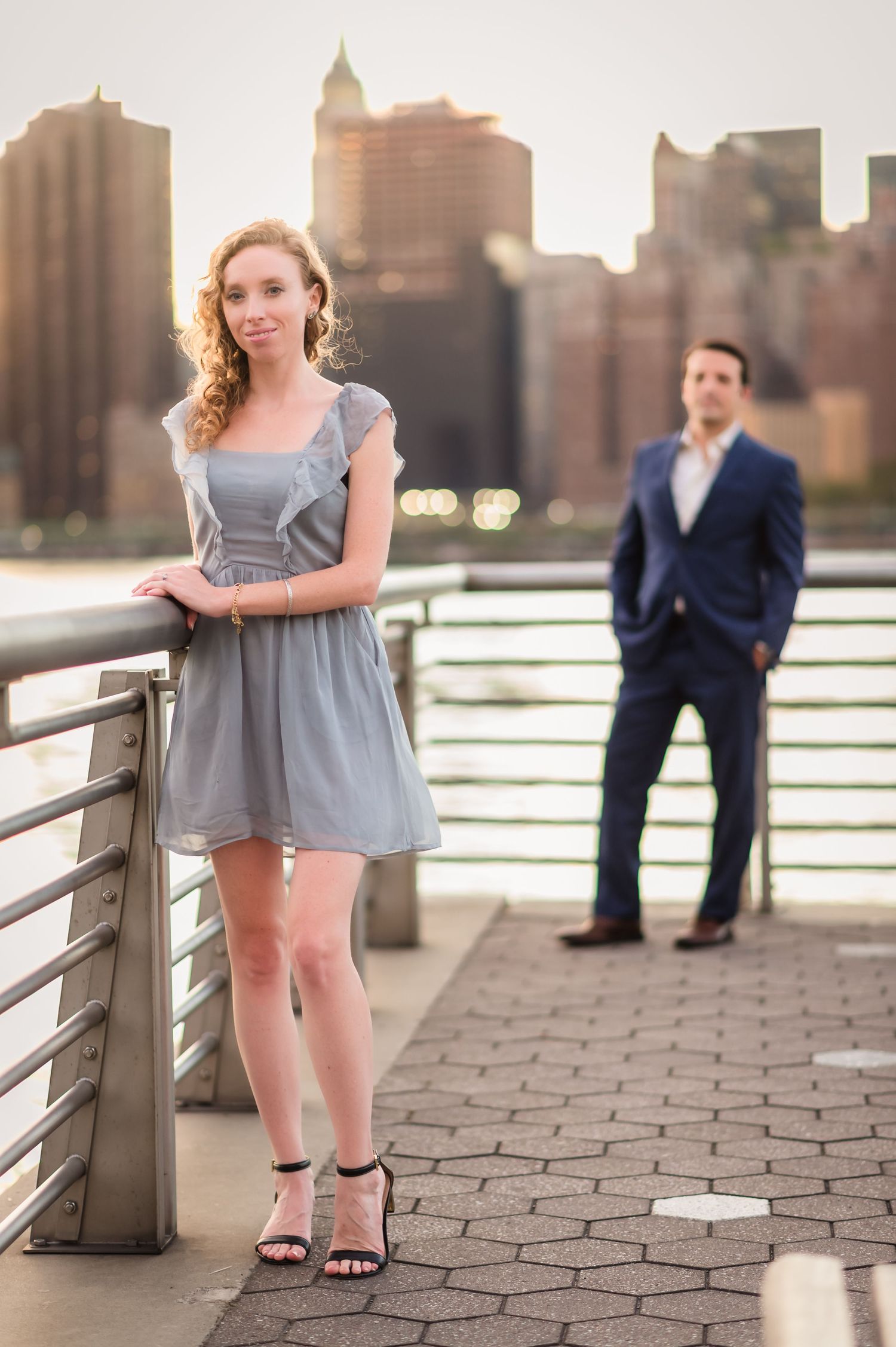 Stylish couple poses along the waterfront with New York City skyline in background.