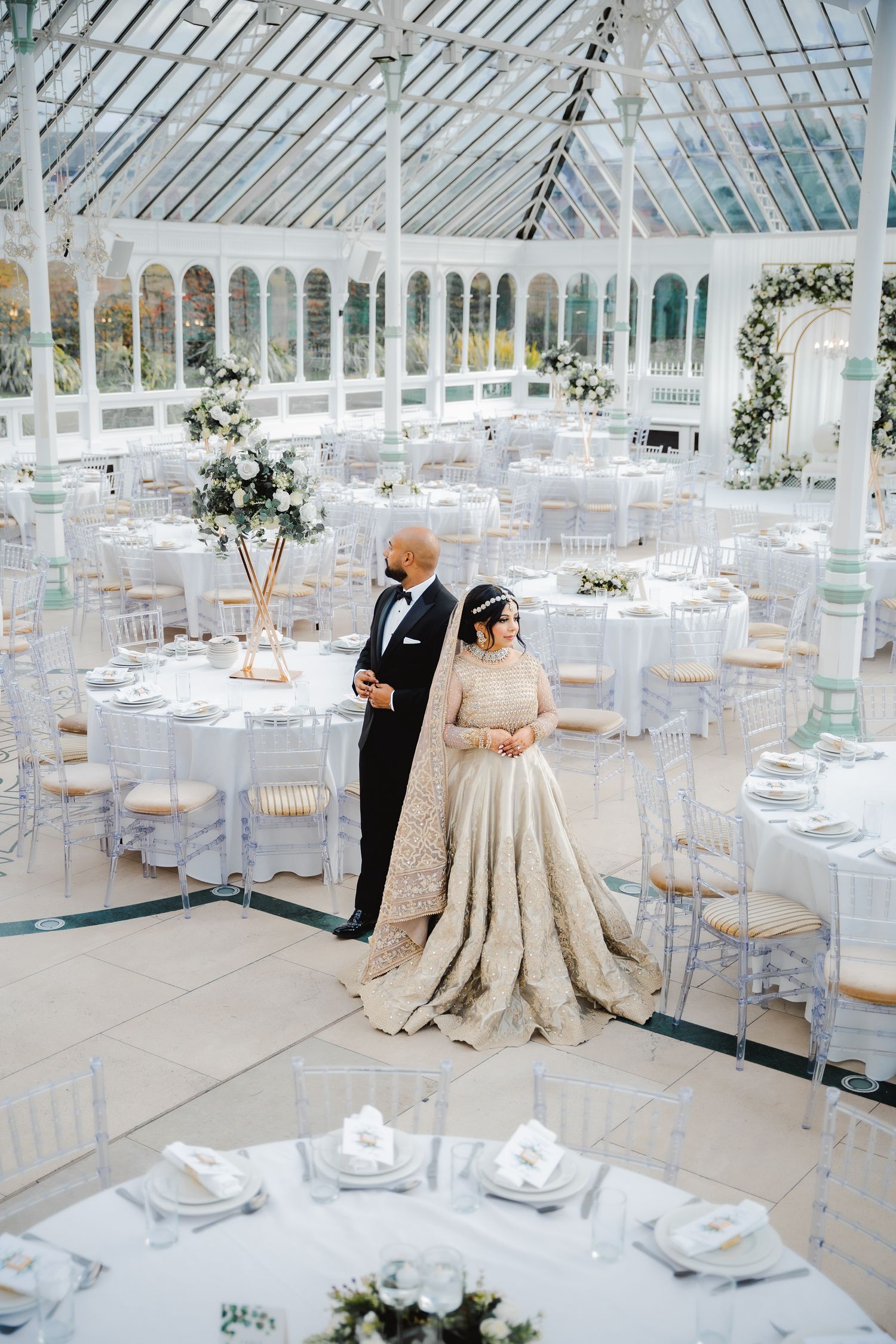 Elegant wedding reception in bright conservatory with white table settings and floral decorations.