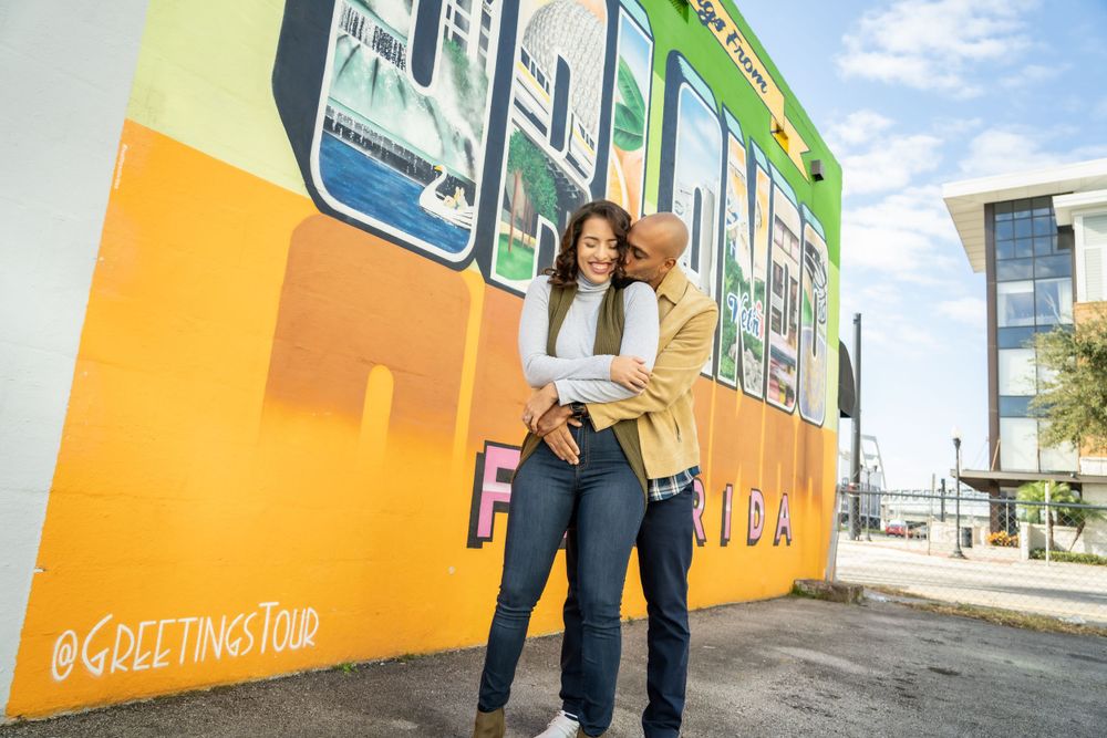 A couple embraces in front of a colorful mural wall outdoors.