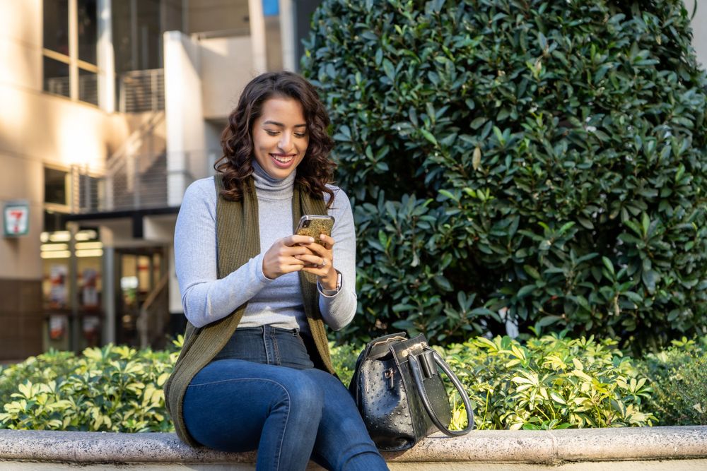 Person in grey sweater using smartphone while sitting outdoors near greenery.