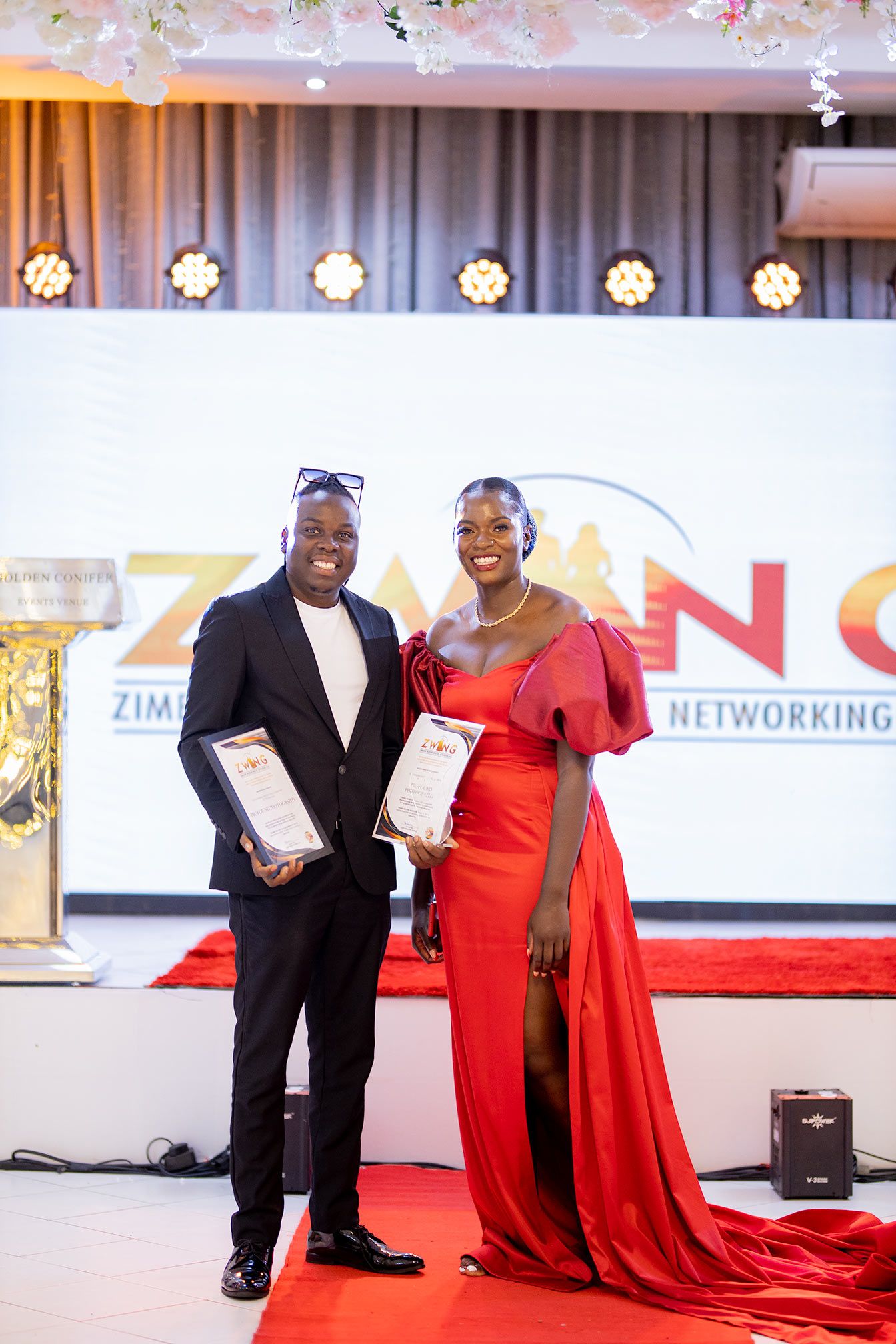 Awards ceremony with two presenters holding certificates on red carpet stage.