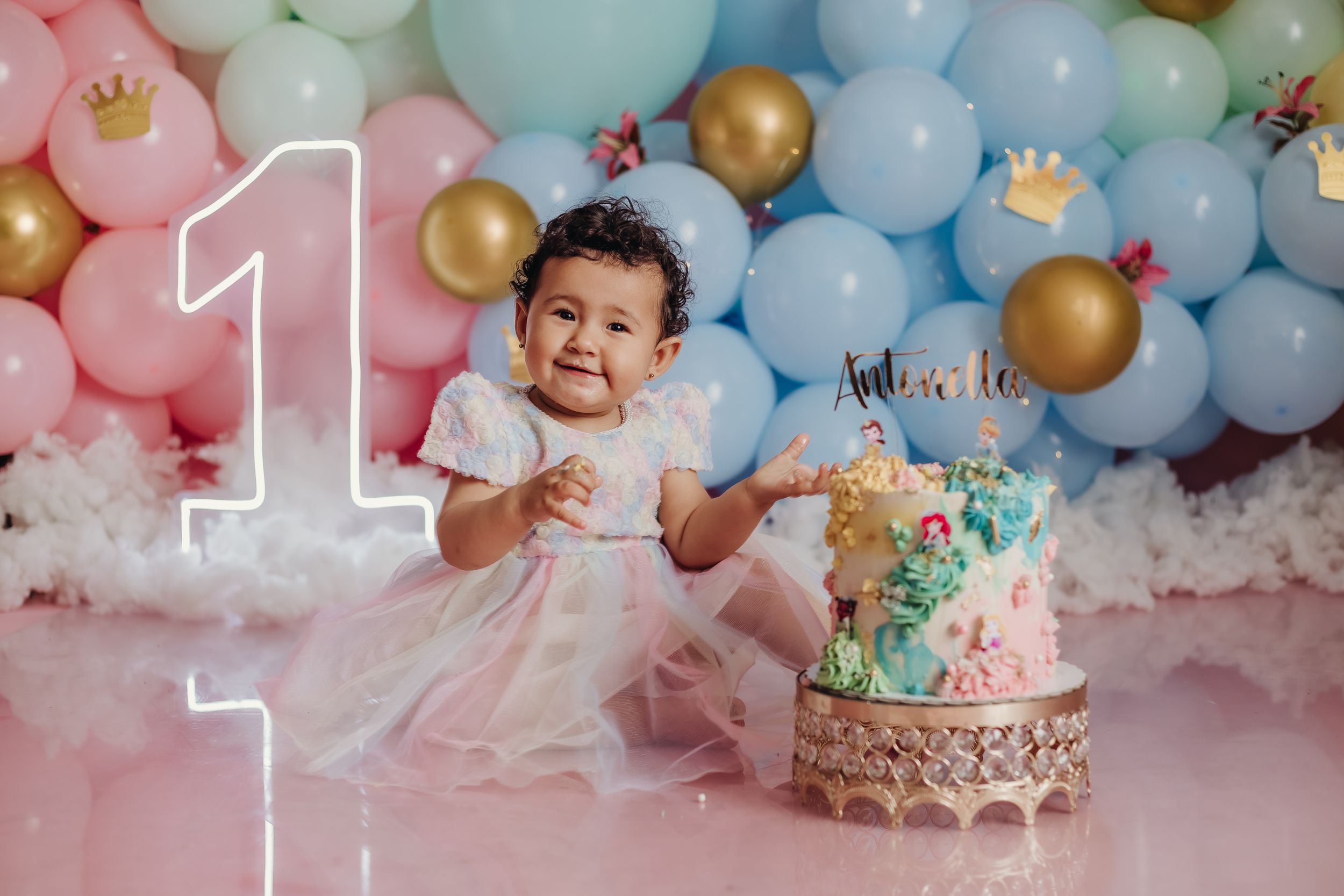 A first birthday celebration setup with pastel balloon arch and decorative cake on a pink backdrop.