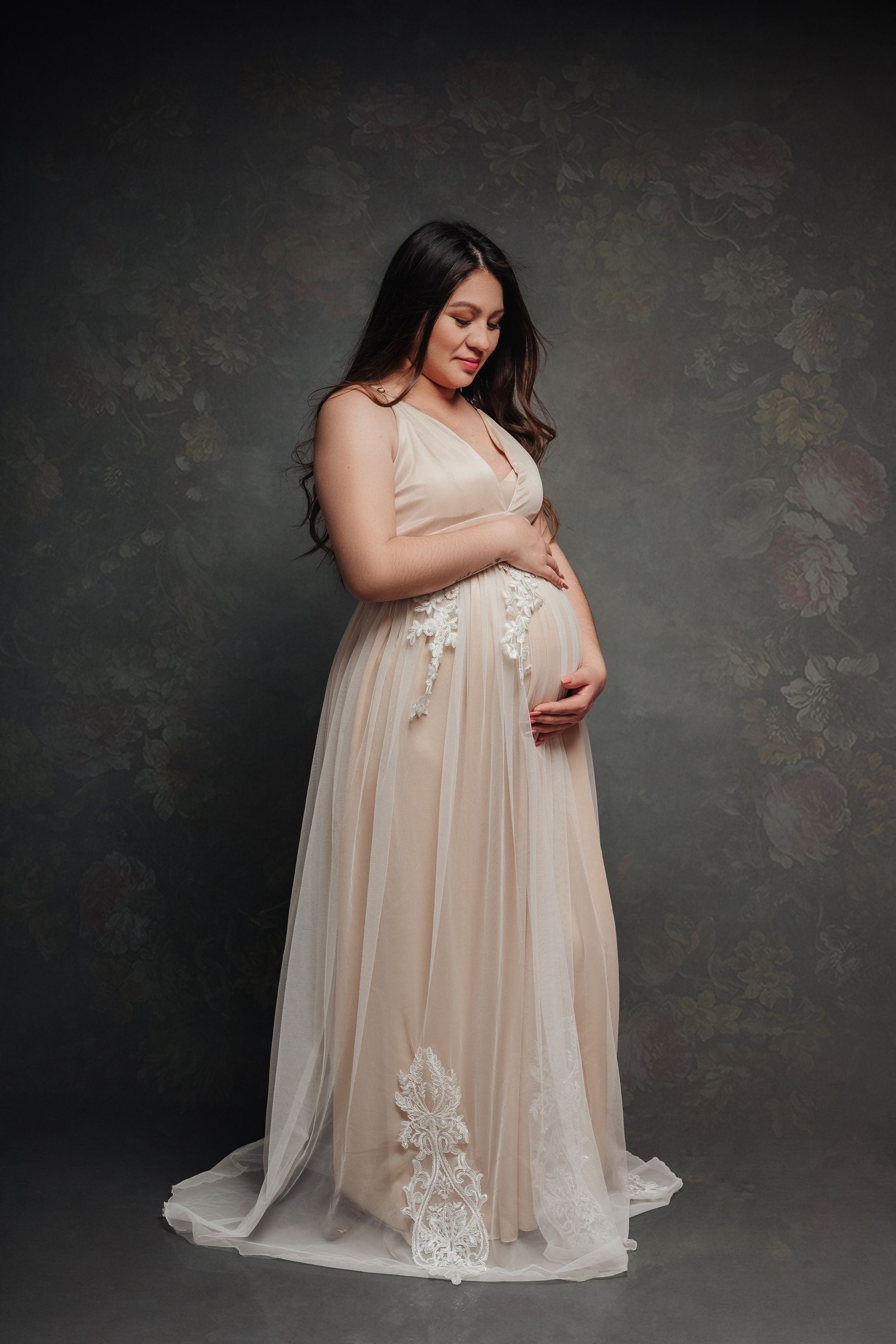 Elegant maternity portrait of woman in flowing champagne gown against dark backdrop.