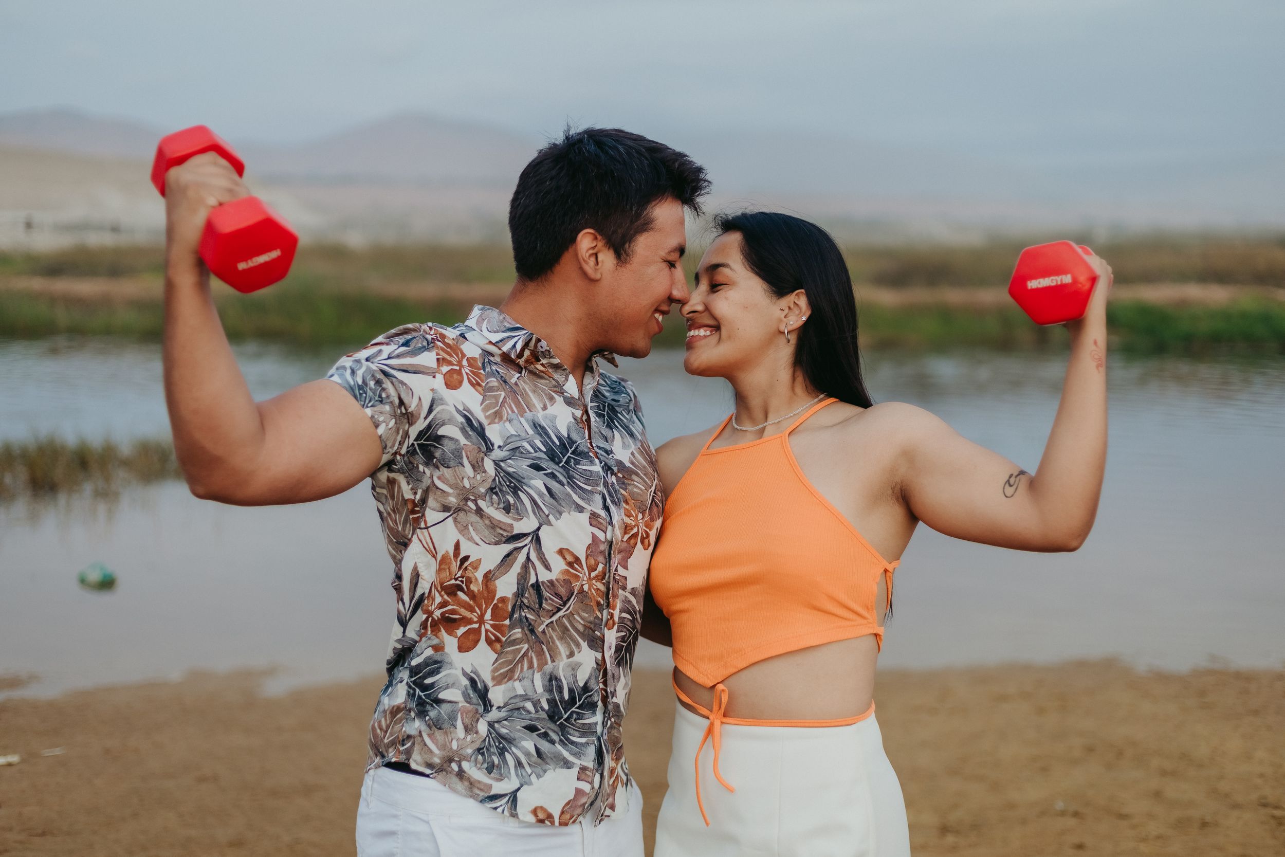 Athletic couple in stylish outfits flexing with red dumbbells outdoors by a lake.