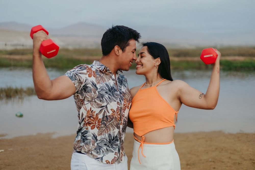 Athletic couple in stylish outfits flexing with red dumbbells outdoors by a lake.