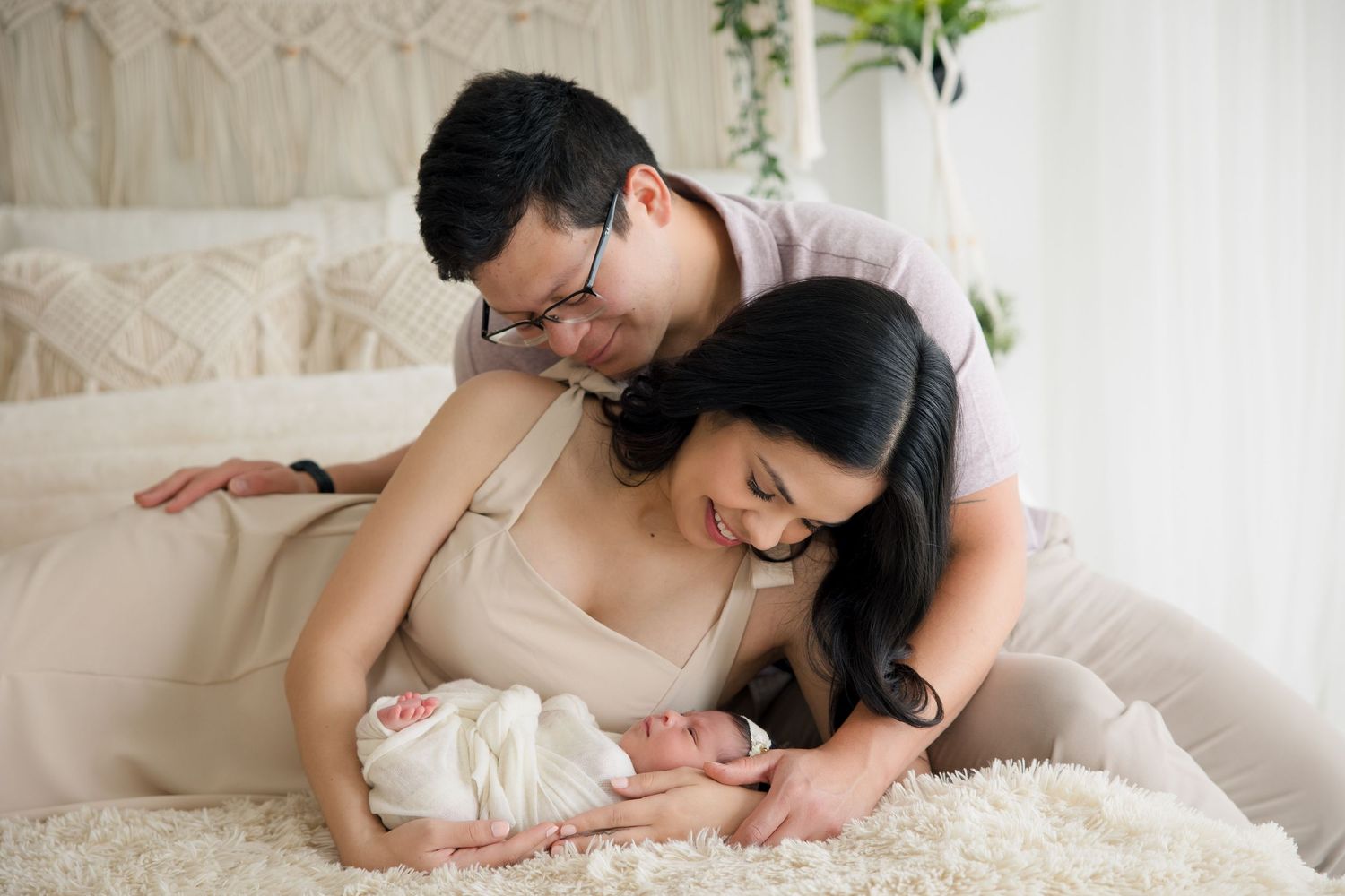 Parents cuddling with their newborn baby on a soft white bed with macrame decor in background.